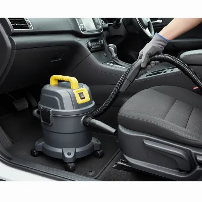 A compact portable shop vacuum with a flexible hose being used to clean the interior of a car, focusing on the carpet and seat crevices. The vacuum is small, on wheels, and easily maneuvered by a person.
